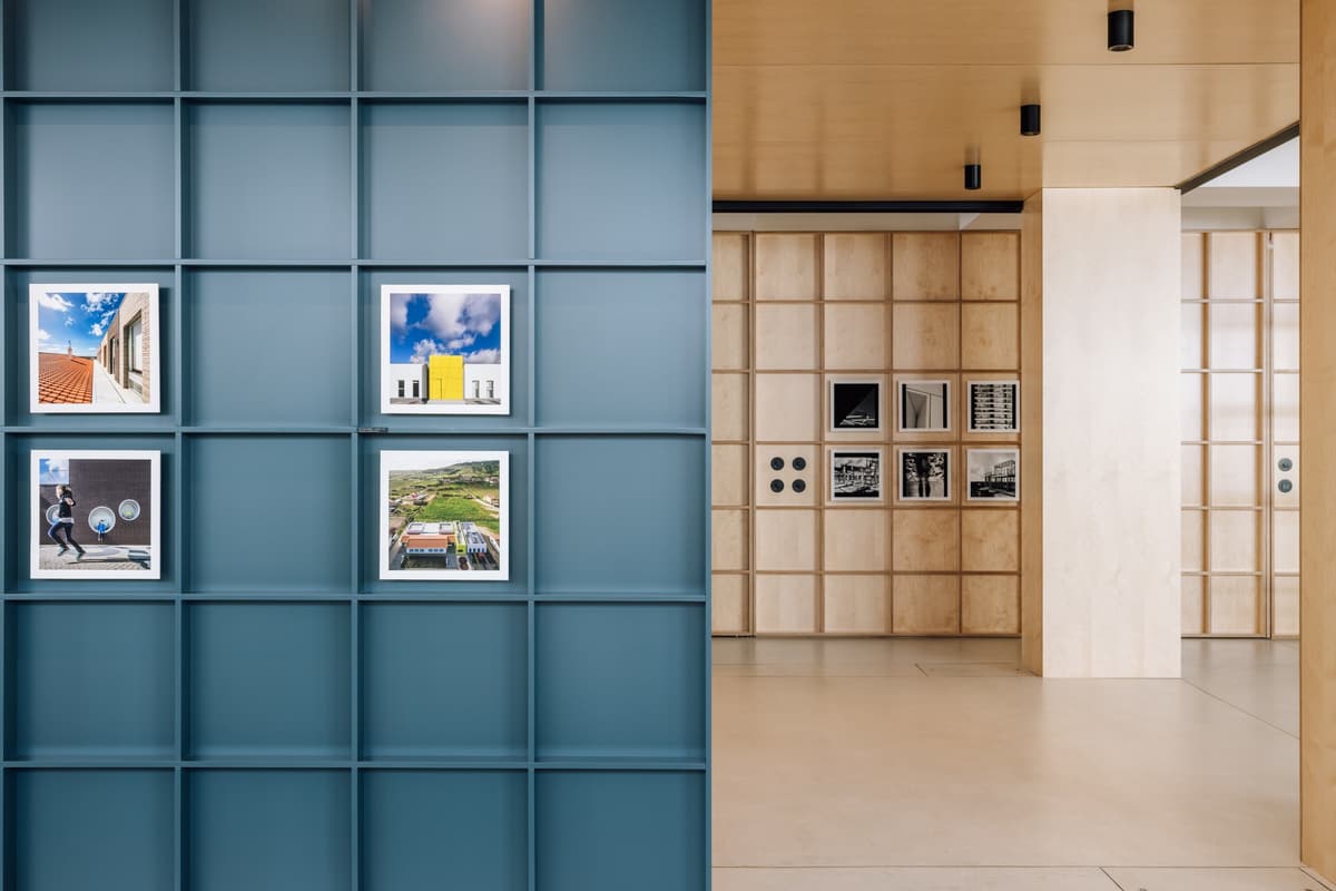 Blue grid wall displaying project photographs, with plywood panelling and a gallery corridor beyond