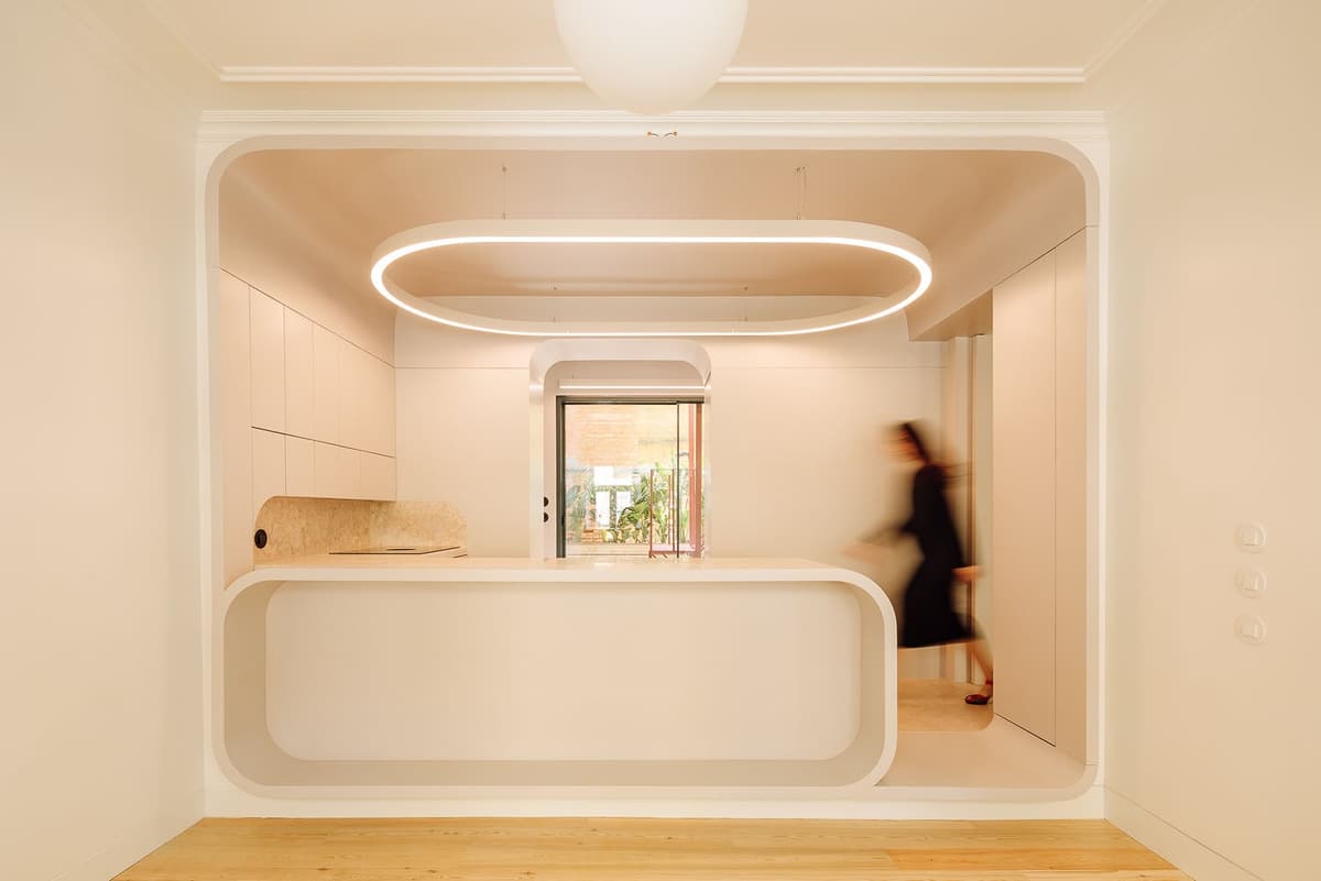 Kitchen in the Colónias flat framed by a rounded white box with an oval light ring, a person walking past