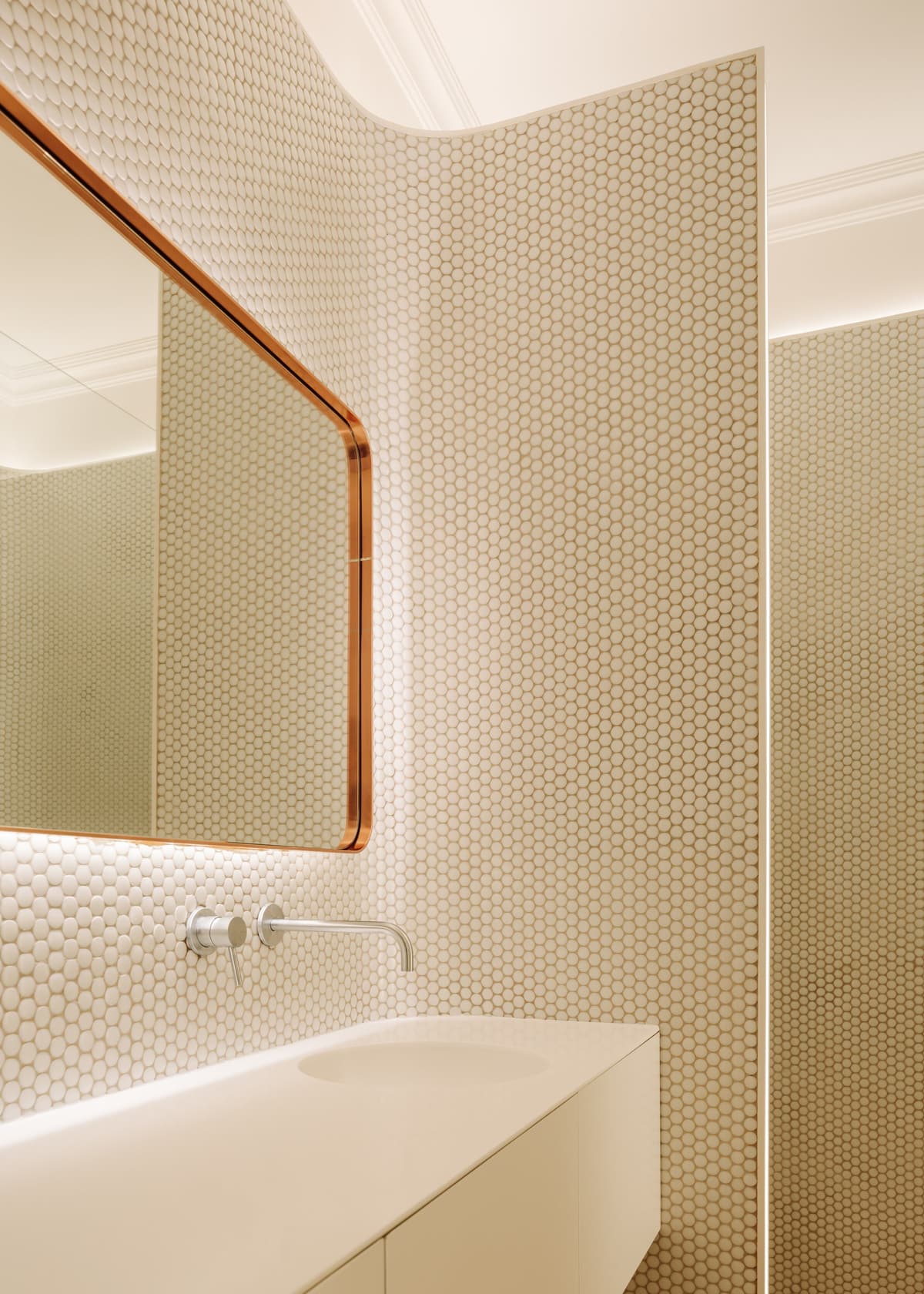 Bathroom with white penny mosaic tiles on a curved wall, copper-framed mirror and white basin