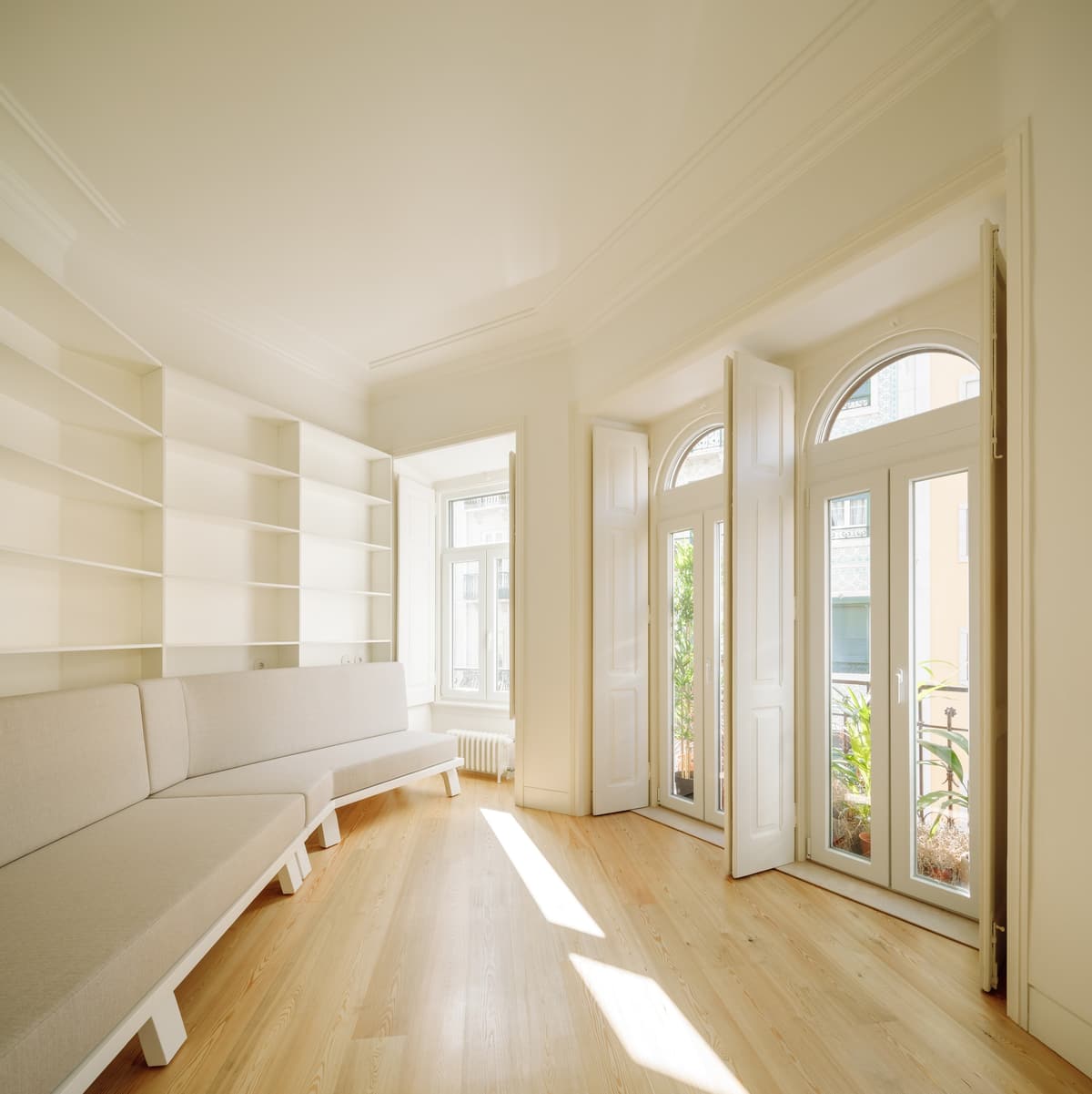 Sunlit living room with built-in white shelves, long white sofa, pine floor and arched balcony doors