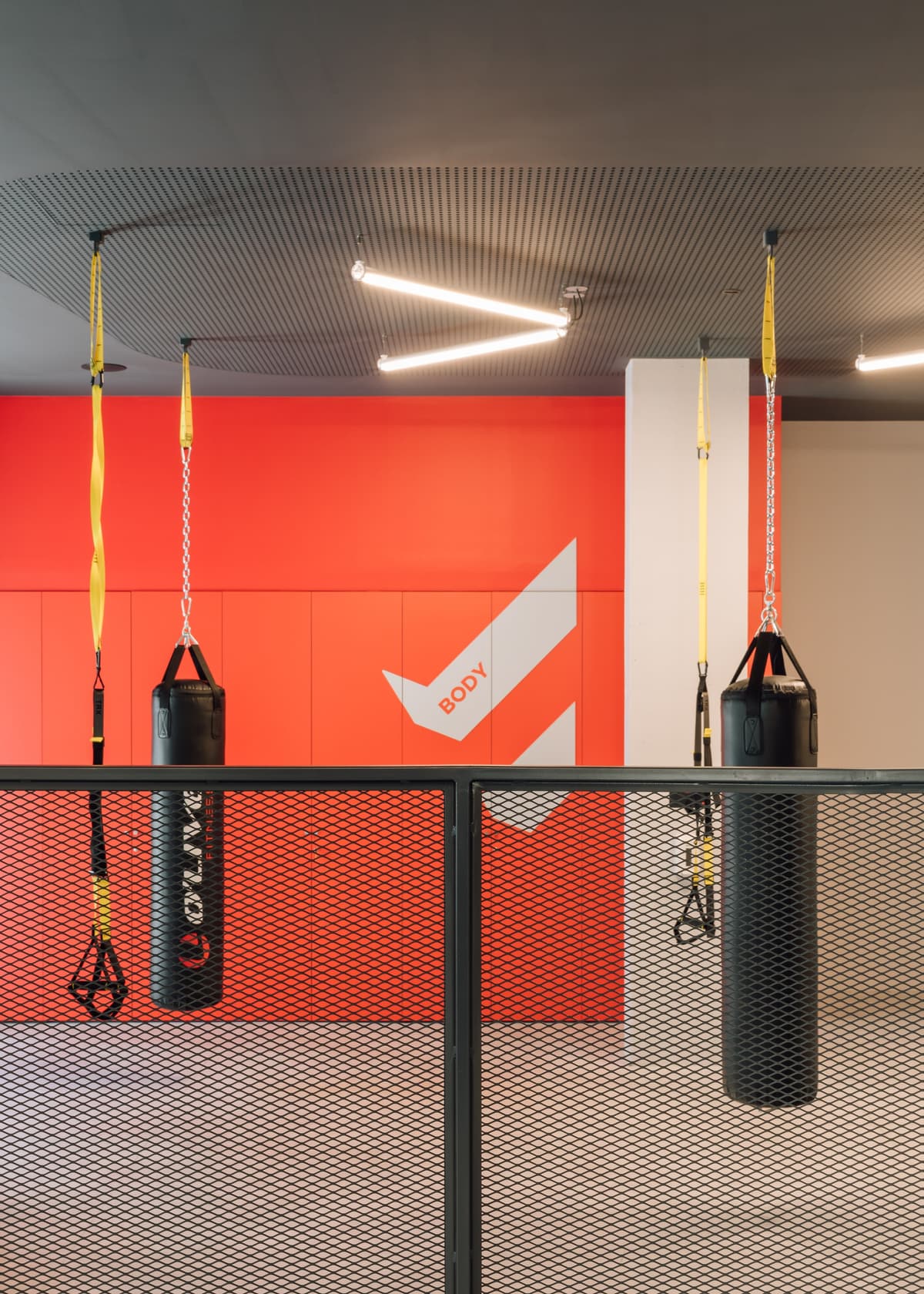 Punching bags and yellow suspension straps behind a black mesh railing, with a red wall marked BODY