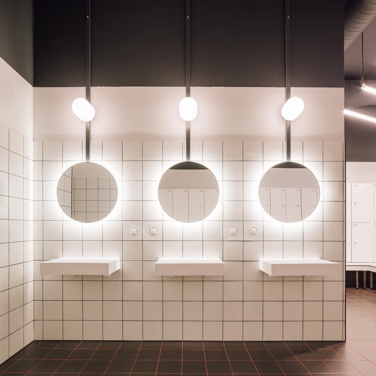 Changing room washbasins with round backlit mirrors, globe lights, white square tiles and dark floor