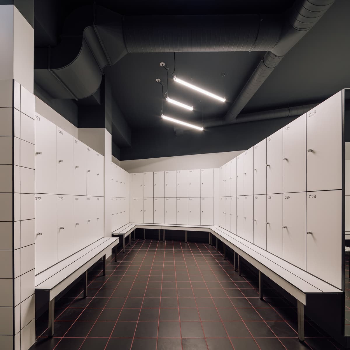 Changing room with numbered white lockers, long benches, exposed ducts and black tiles with red grout