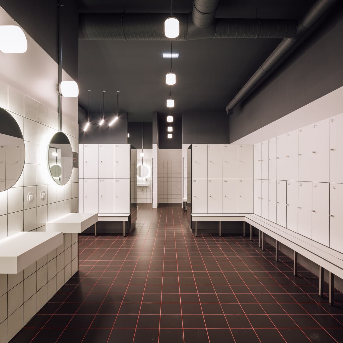 Changing room with backlit round mirrors, white lockers and benches on a black and red tiled floor