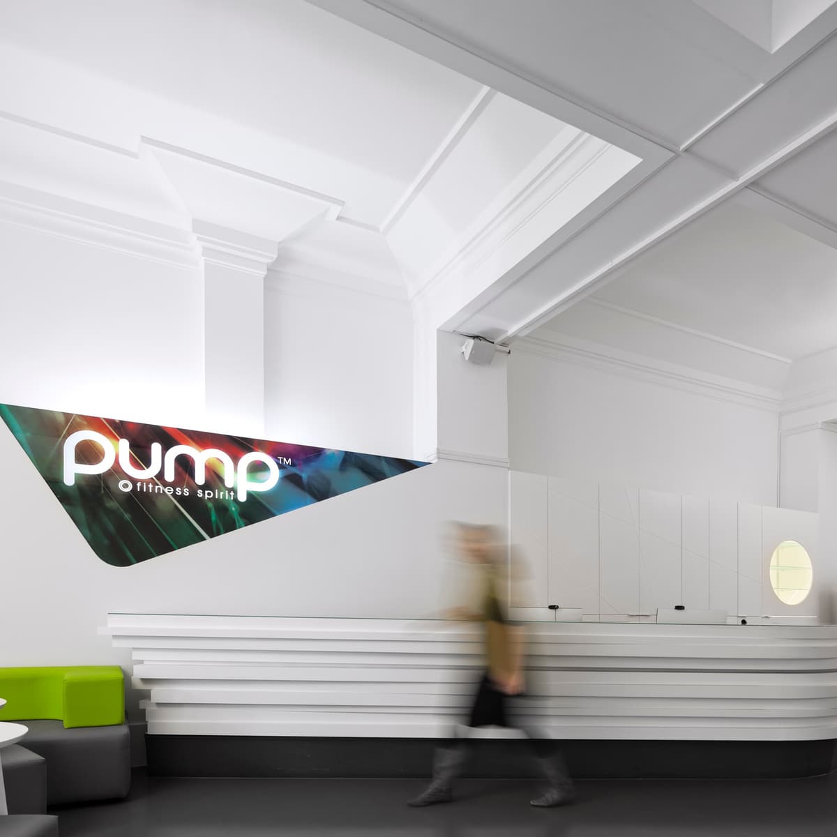 Pump gym reception with a layered white counter, illuminated logo panel and a visitor walking past in motion blur
