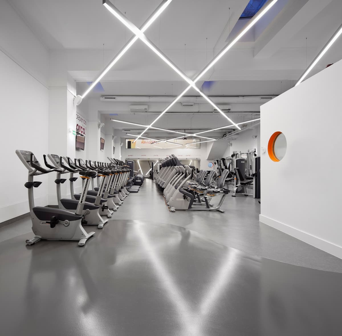 Gym floor with rows of exercise bikes under crossing linear lights, white walls and an orange-rimmed round window