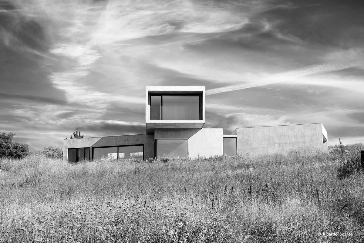 Black and white view of the Adão Lobo house, a cantilevered volume rising above low blocks in tall grass