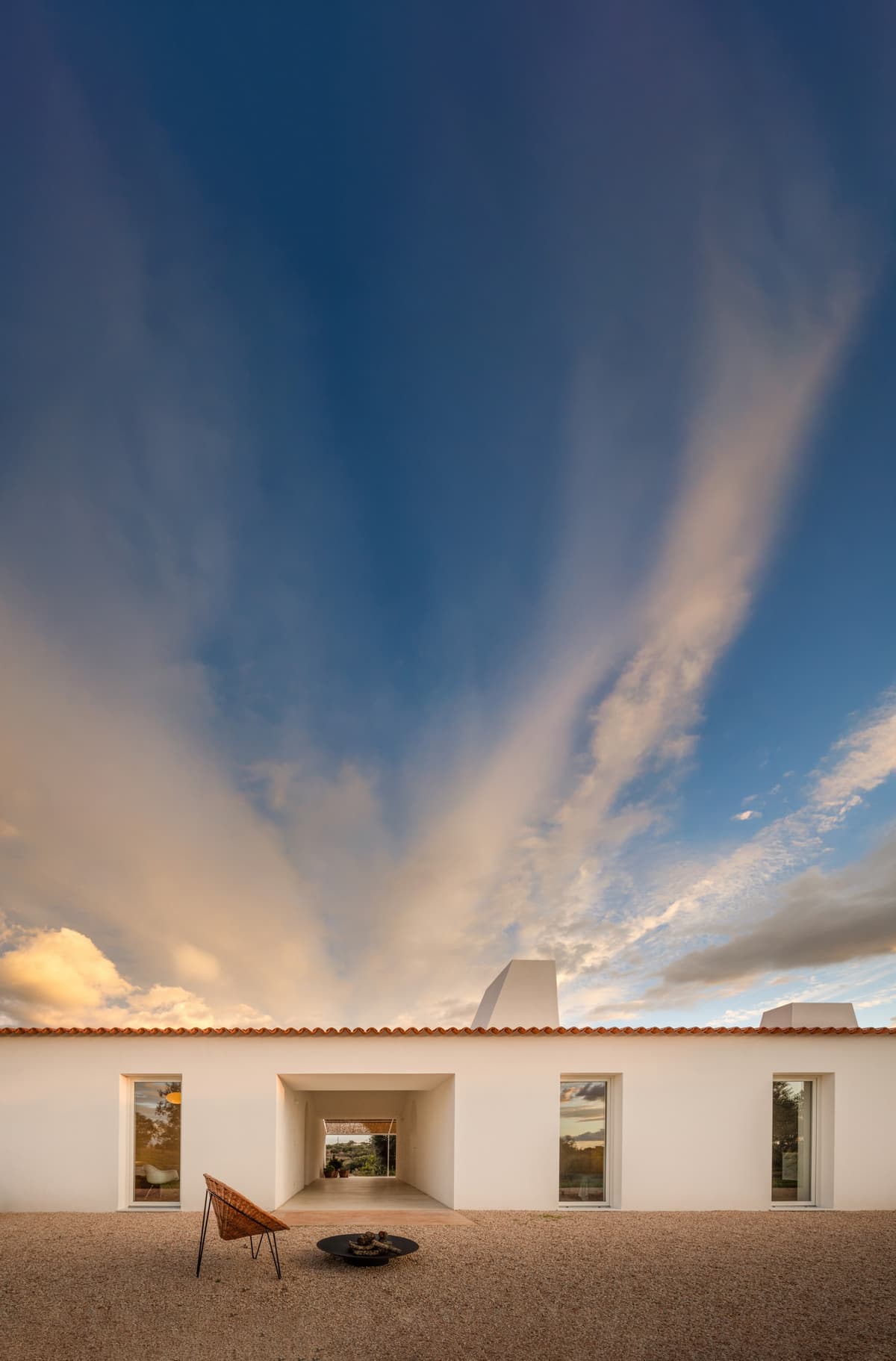 Whitewashed facade of Monte Alvo with a deep central passage and chimneys, wicker chair on gravel at dusk