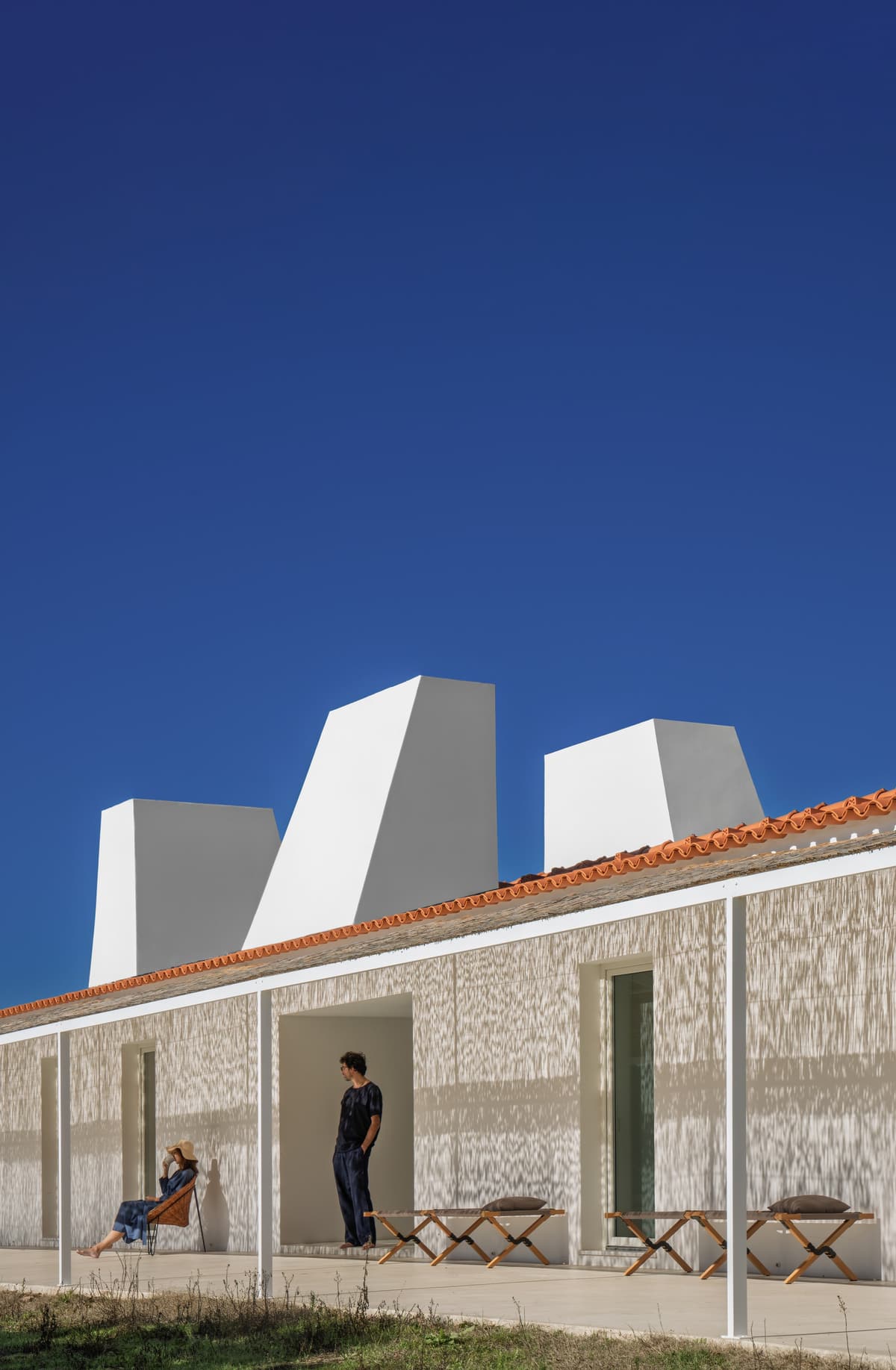 Sculptural white chimneys above a tiled roof and shaded porch, two people resting against the sunlit wall