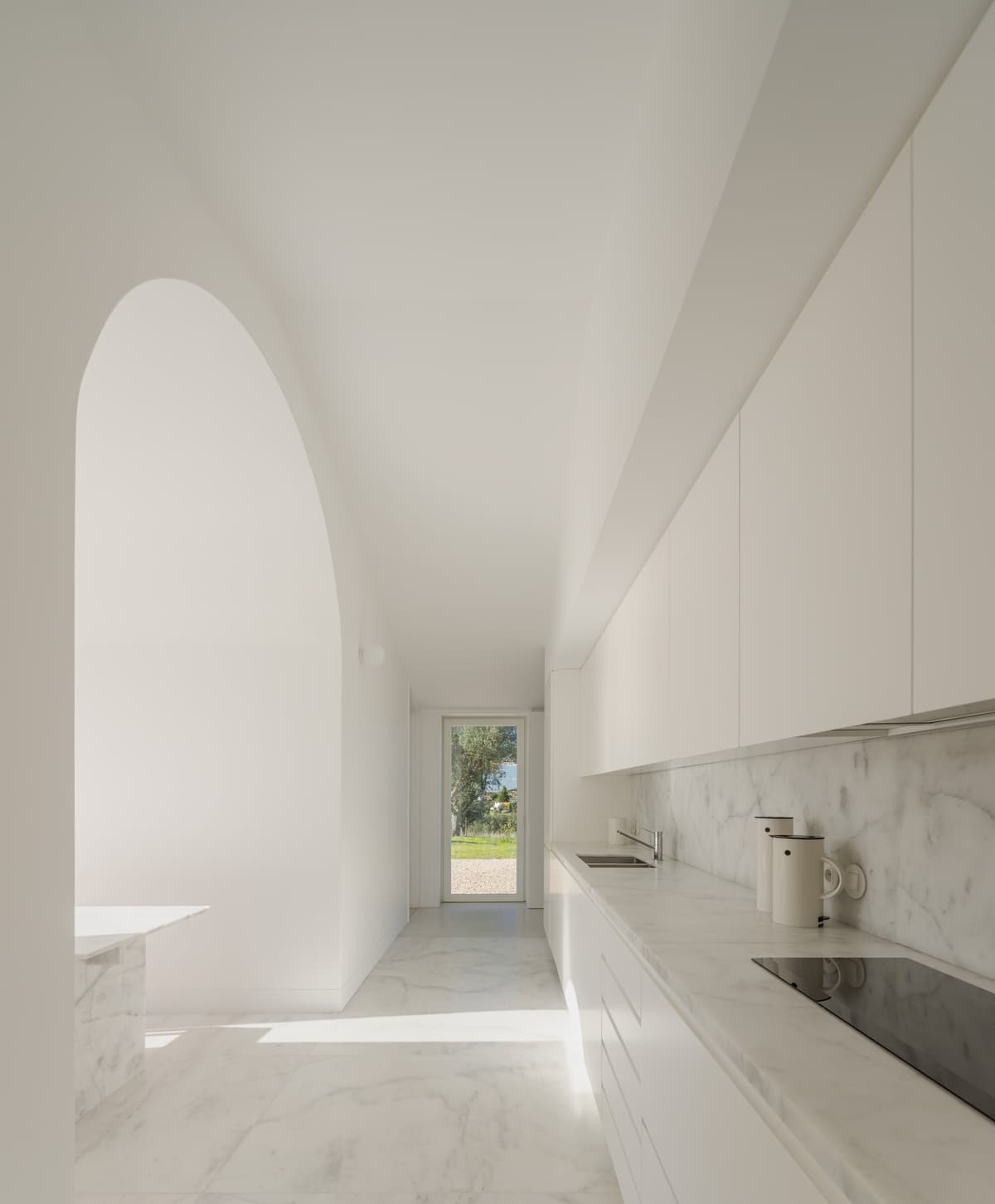 White kitchen with marble worktops and floor, a tall arched opening and a glazed door to the garden
