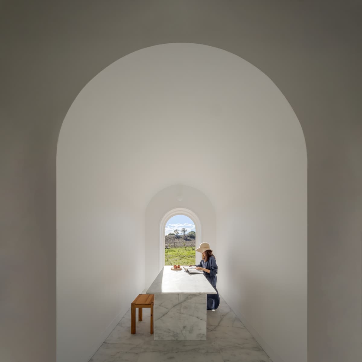 Vaulted white room with a marble table, a person reading beside an arched window framing the fields