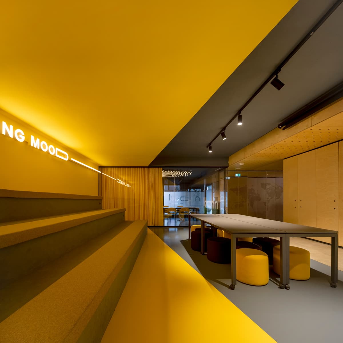 Yellow tiered seating with illuminated lettering beside grey tables and round stools in the Nhood Lisbon office