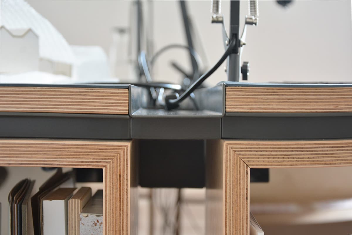 Detail of a plywood desk with dark linoleum top and a central metal cable gutter for plugs