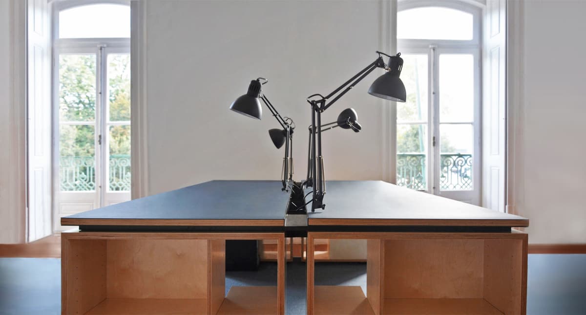 NOZ studio desk in plywood with dark linoleum top and black task lamps, tall balcony windows behind