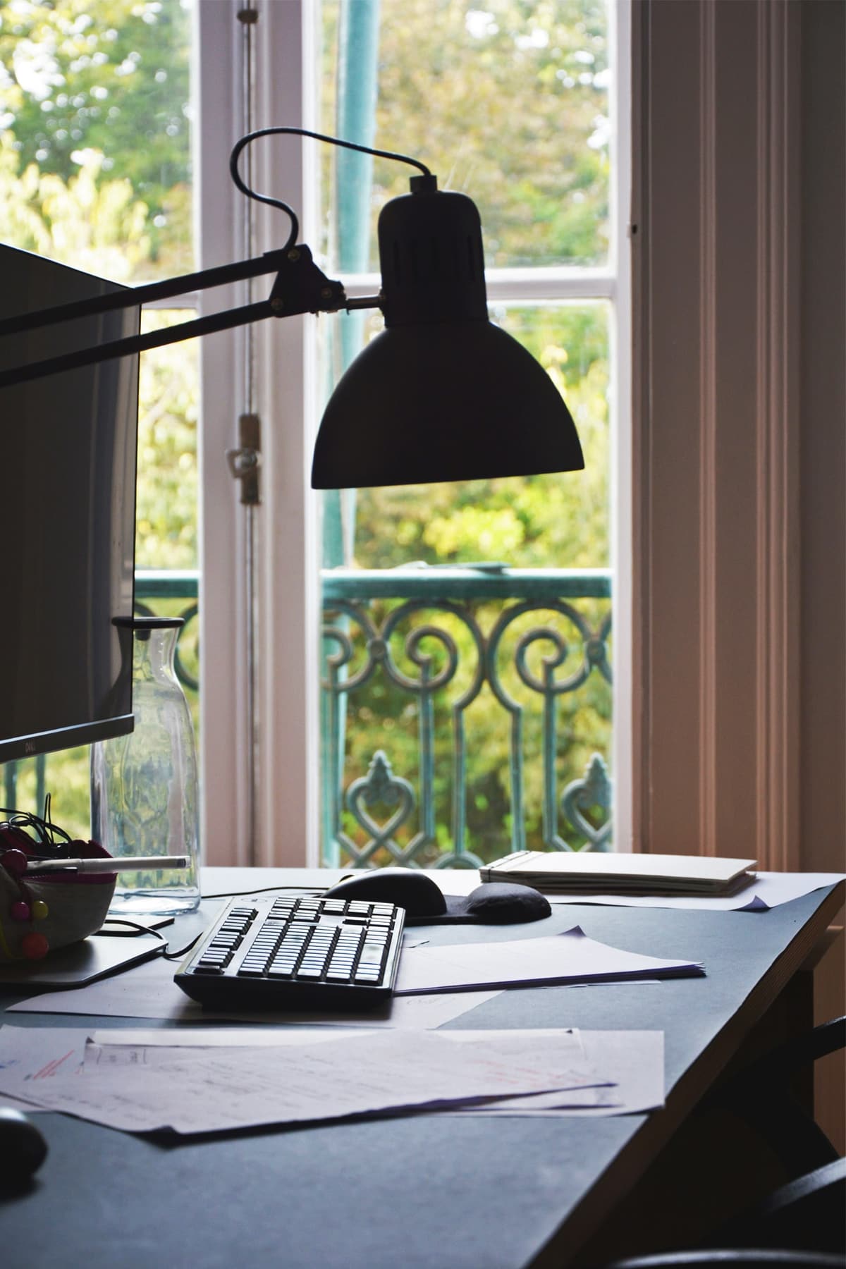 Workstation with monitor, keyboard and black desk lamp by a tall window with wrought-iron balcony and garden