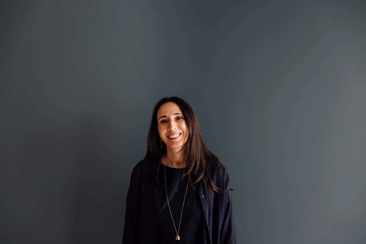 Portrait of inês sousa, in a black jacket and long necklace, against a dark grey wall