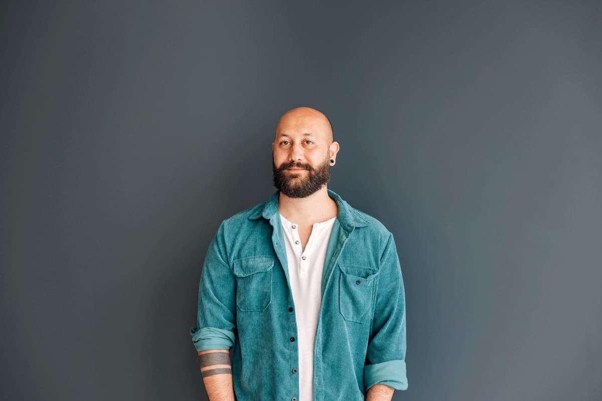 Portrait of josé malhó, in a teal corduroy shirt over a white henley, against a dark grey wall