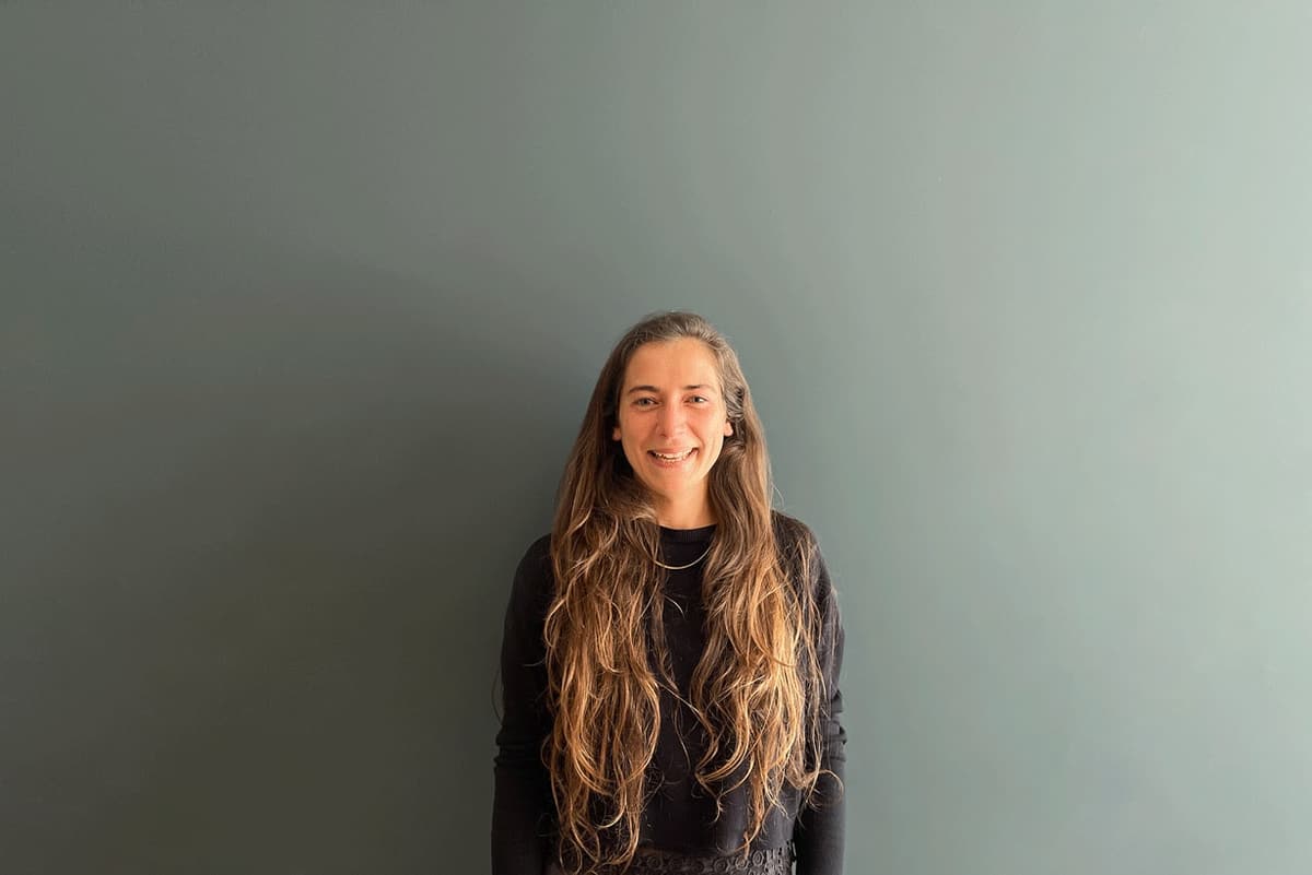 Portrait of maria inês gonçalves, with long wavy hair and a black top, against a grey-green wall