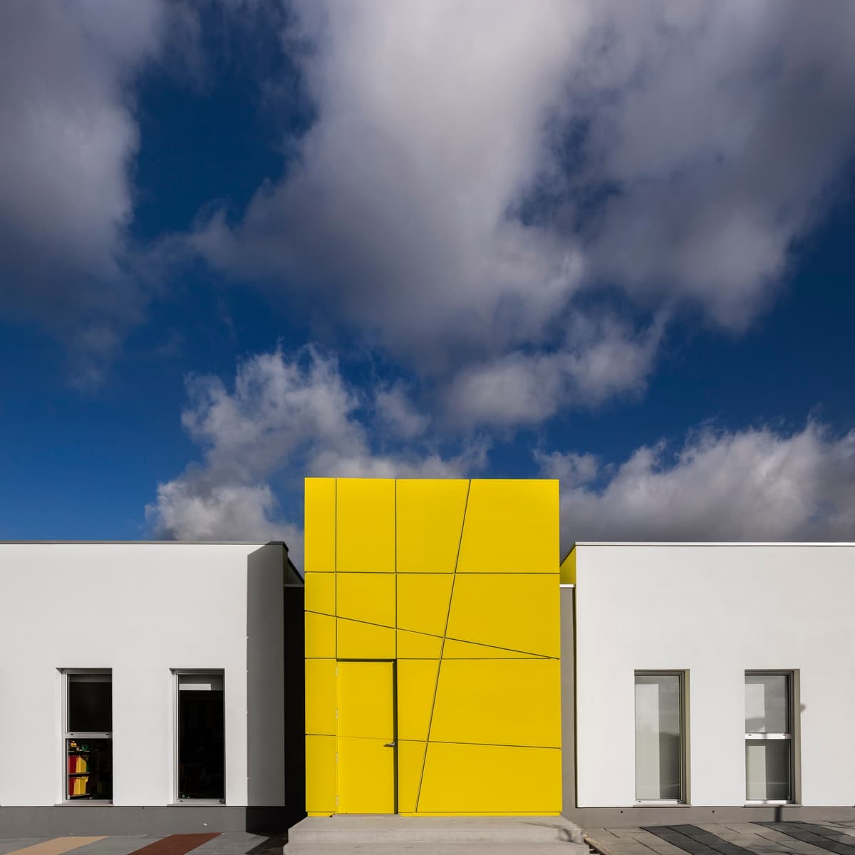 Alguber primary school entrance, a yellow panelled volume between white single-storey wings under a cloudy sky