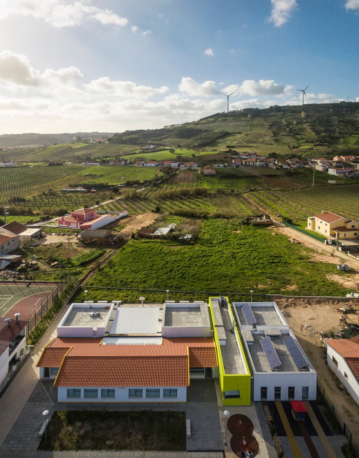 Aerial photograph of Alguber primary school with tiled roofs, green-yellow volume and playground amid farmland