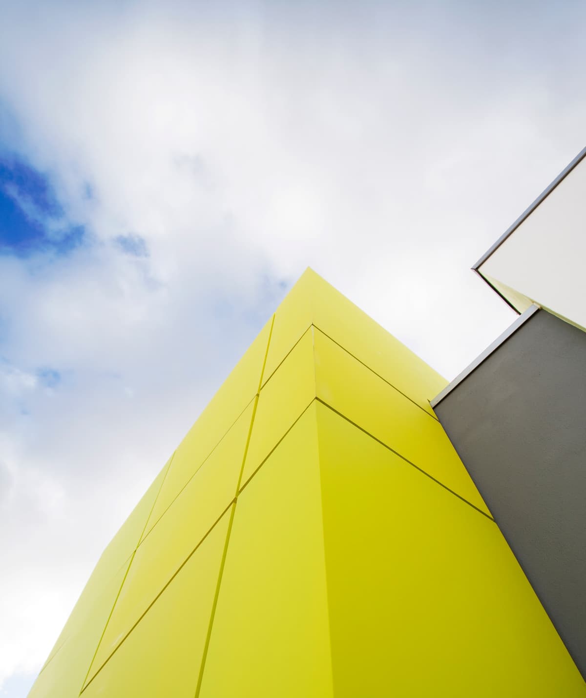 Upward view of the yellow-green panelled entrance volume of Alguber school against a cloudy sky