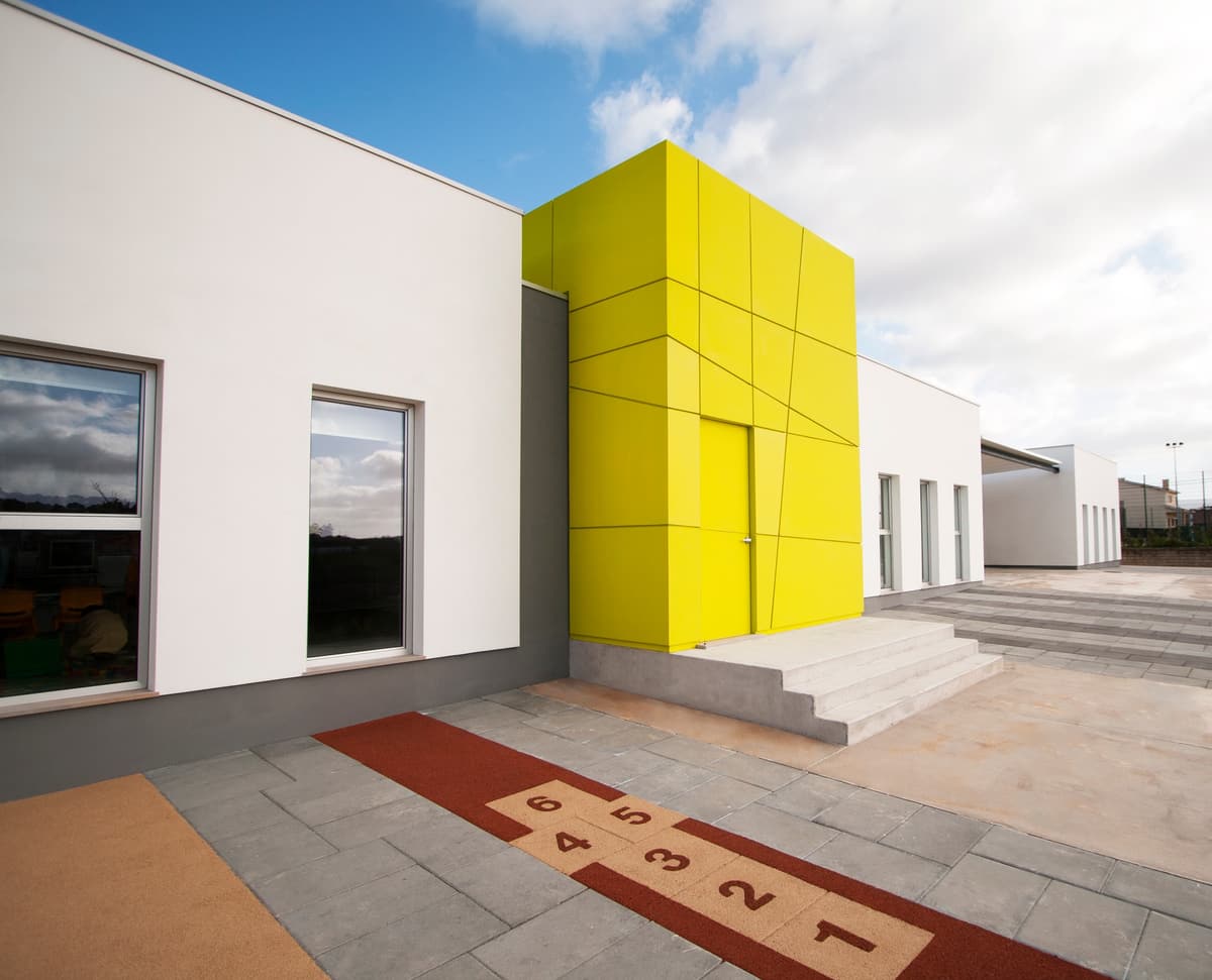 Yellow entrance volume with steps beside white classroom wings, a hopscotch pattern on the playground paving