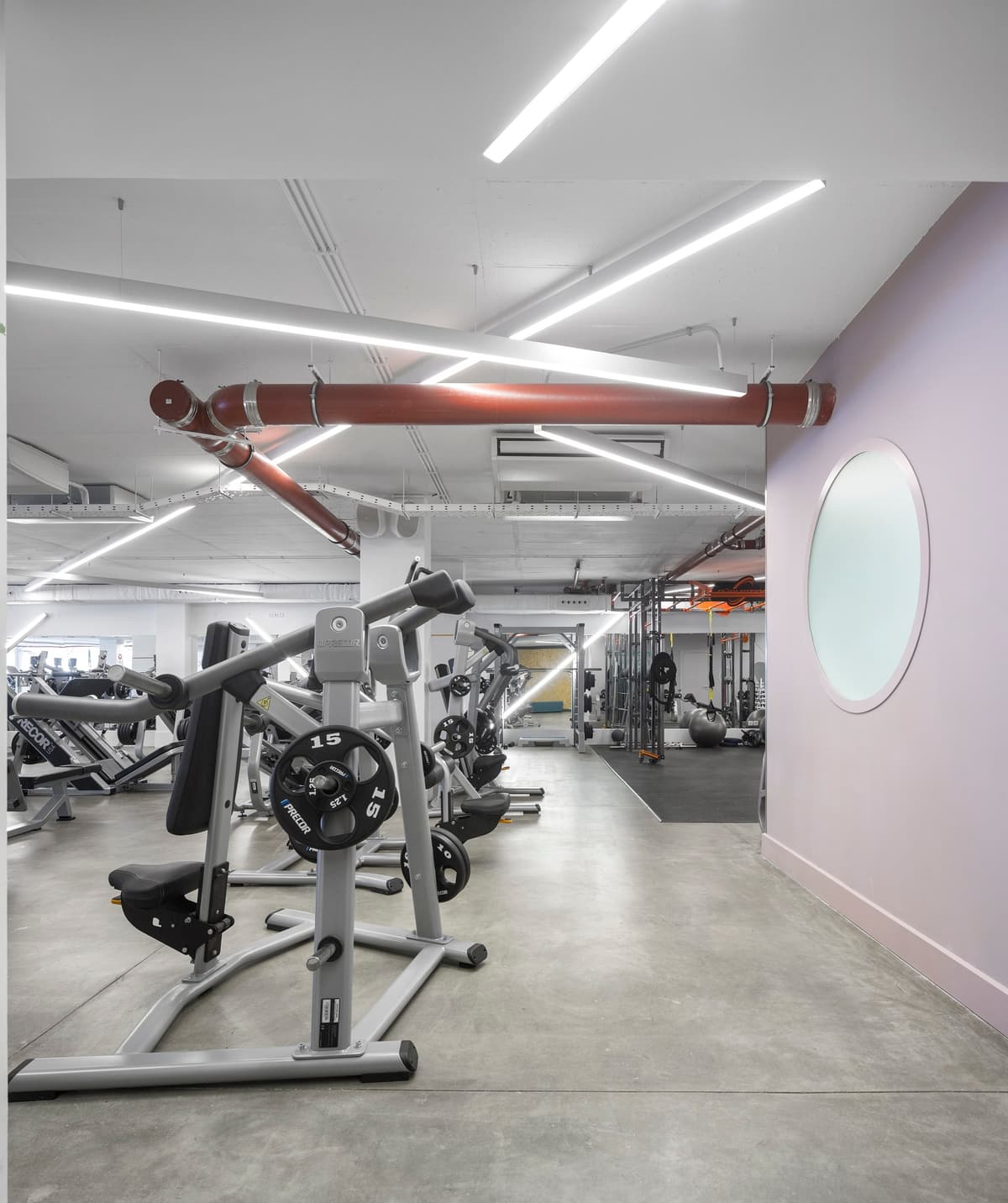 Weight training area at Pump Alvalade with exposed red duct, linear lights and a round window in a lilac wall