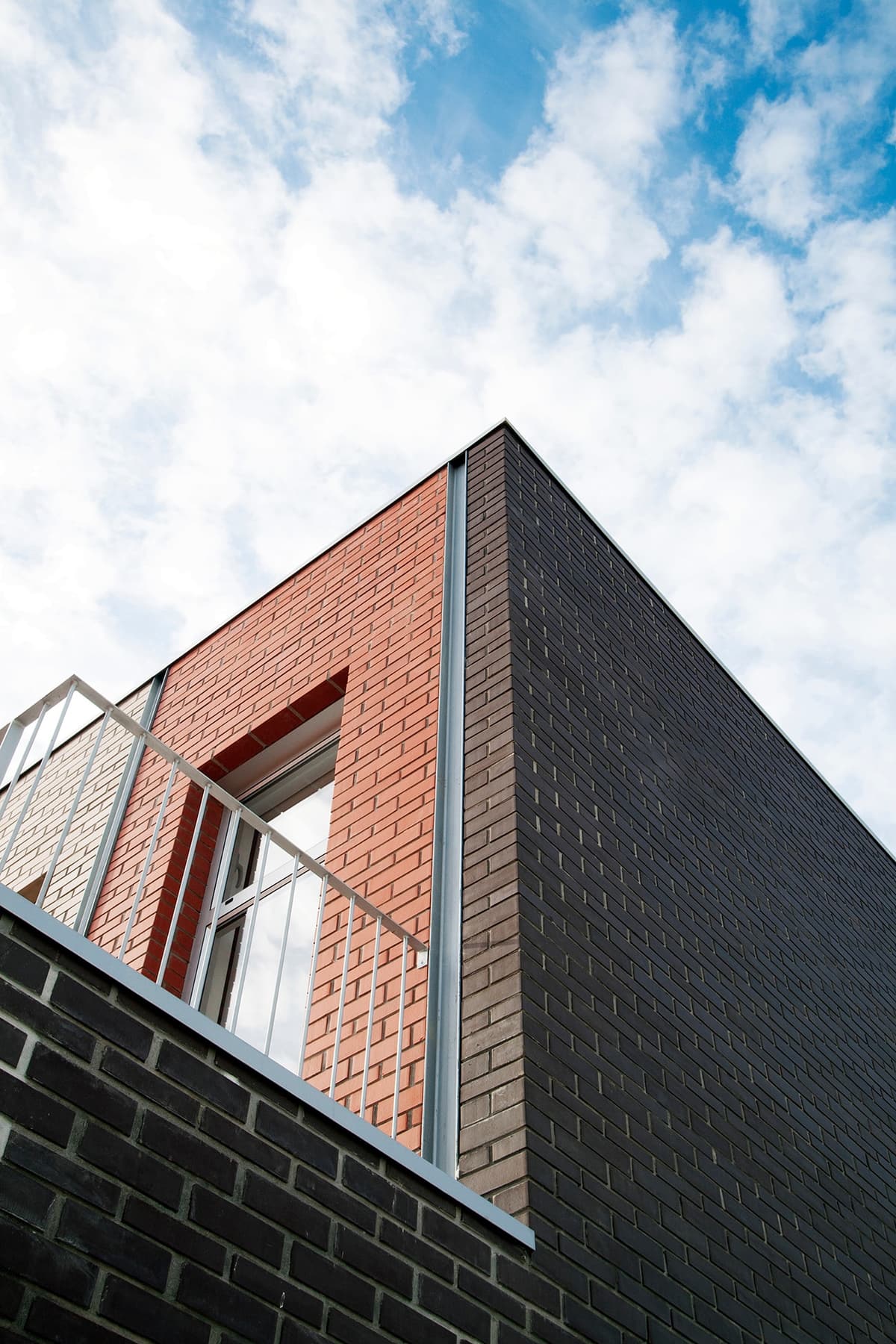 Low-angle view of red and dark brick volumes with a balcony railing against a cloudy sky