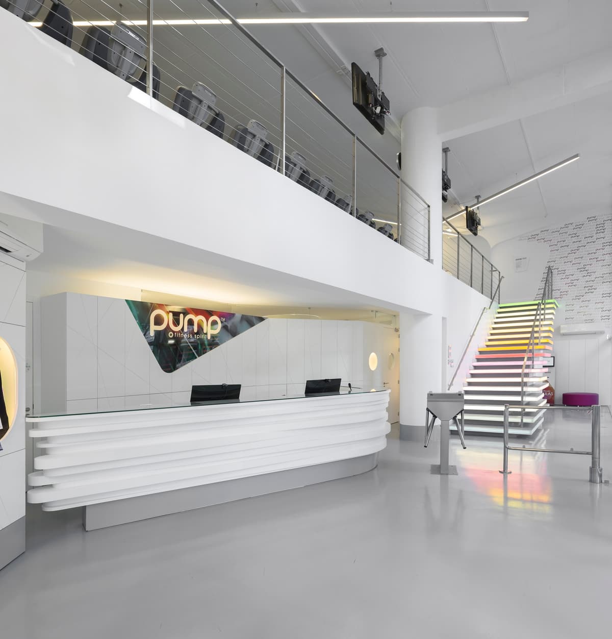 Pump Expo reception with a layered white curved desk, a mezzanine of cardio machines and a lit colour staircase