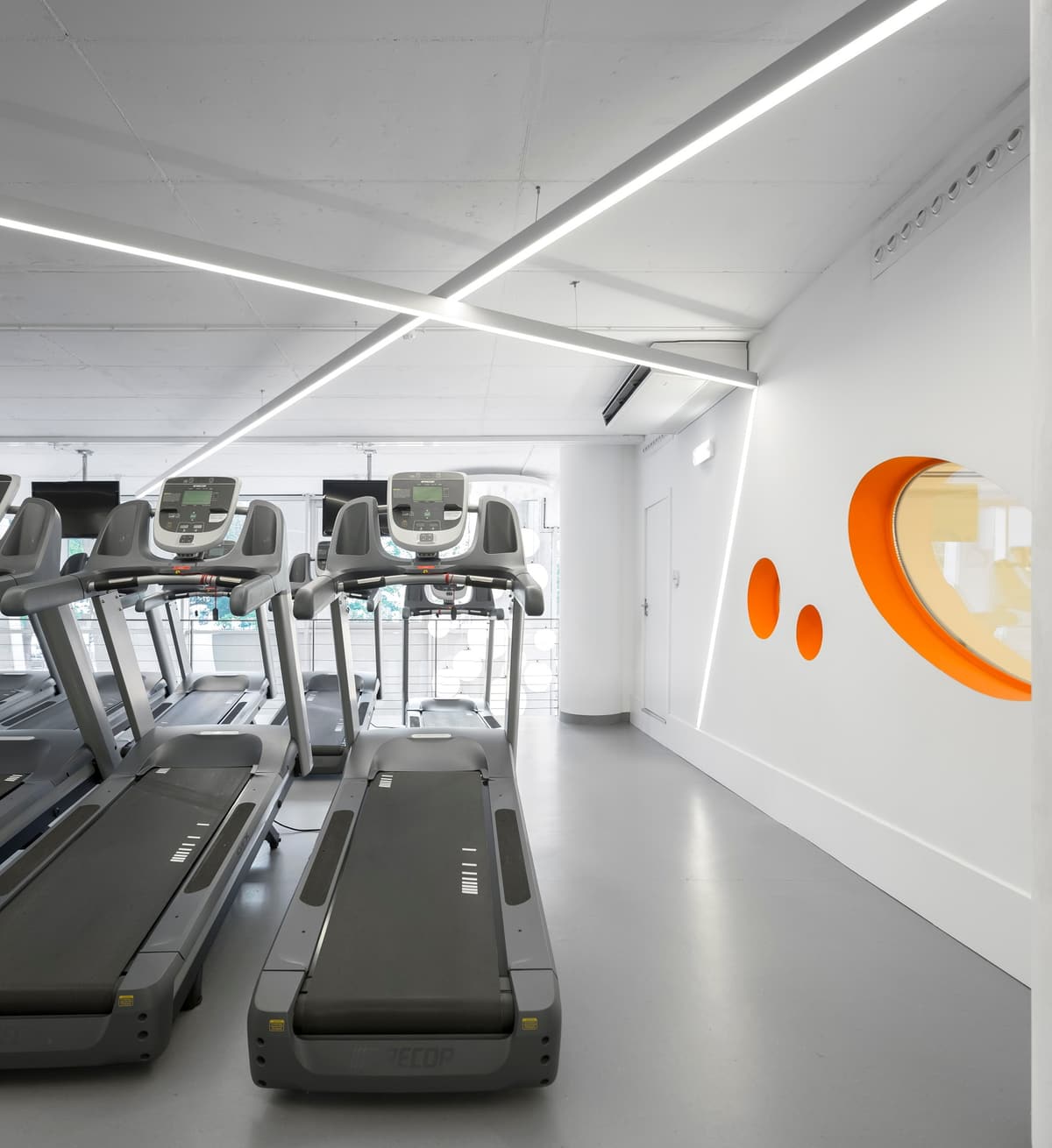 Row of treadmills beside a white wall with round orange-rimmed windows under crossed linear lights, Pump Expo