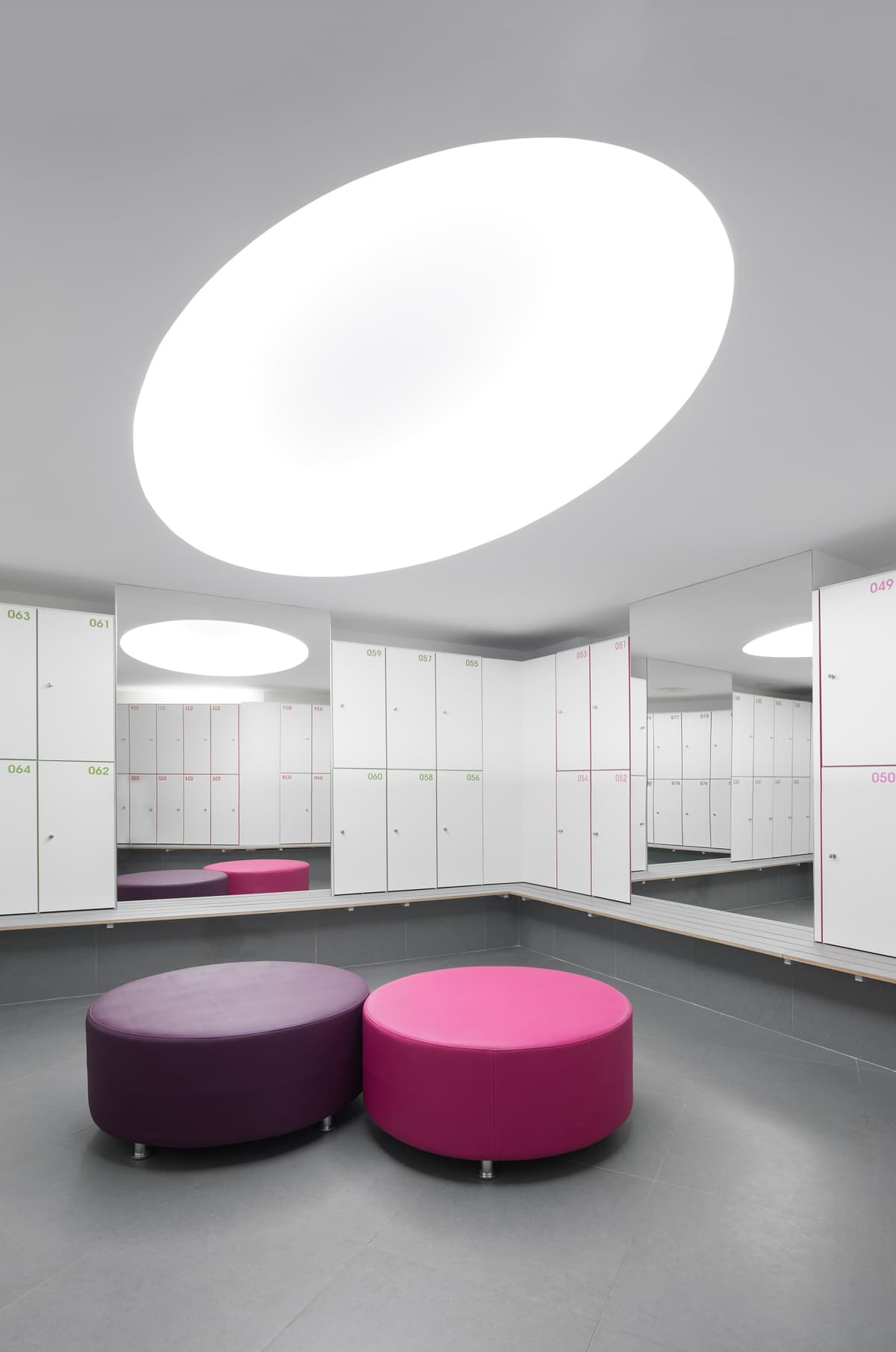 Changing room with white lockers, mirrors, an oval luminous ceiling and round purple and pink seats
